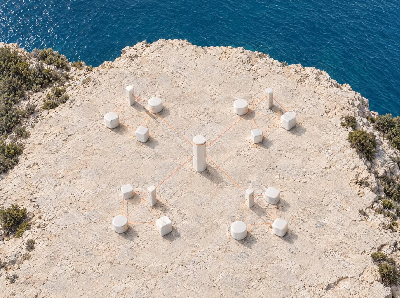 Aerial view of four separate clusters of white modules on a clifftop plaza, each linked by orange filaments to one central pillar.